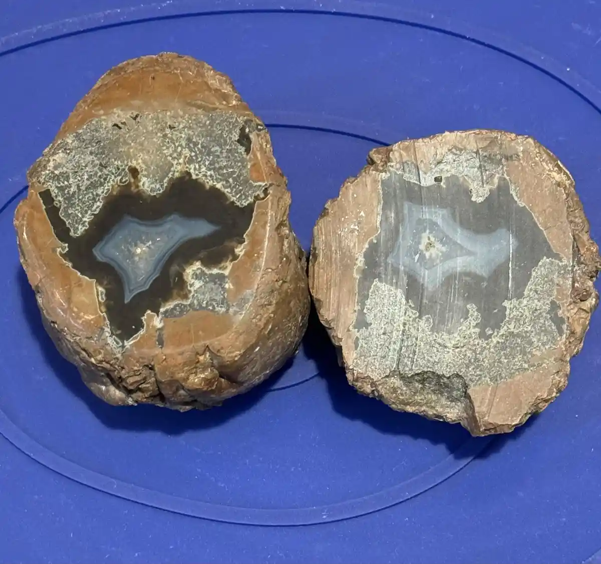 Two halves of a split geode rest on a blue plastic surface, revealing their internal crystal formations. The left half shows…