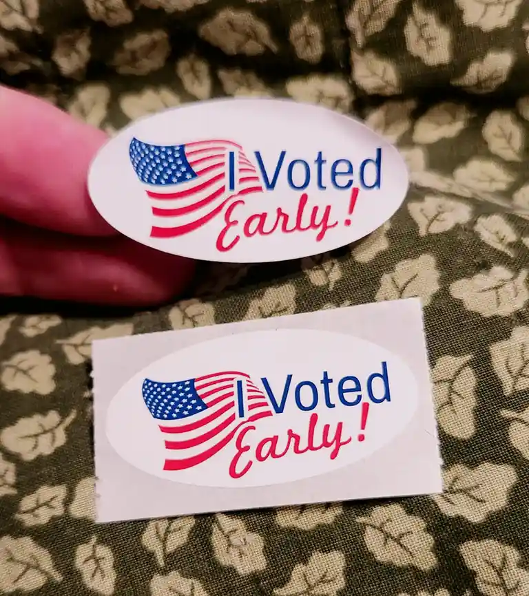 A close-up photo shows a person holding one oval 'I Voted Early!' sticker, while another identical sticker lies on a…