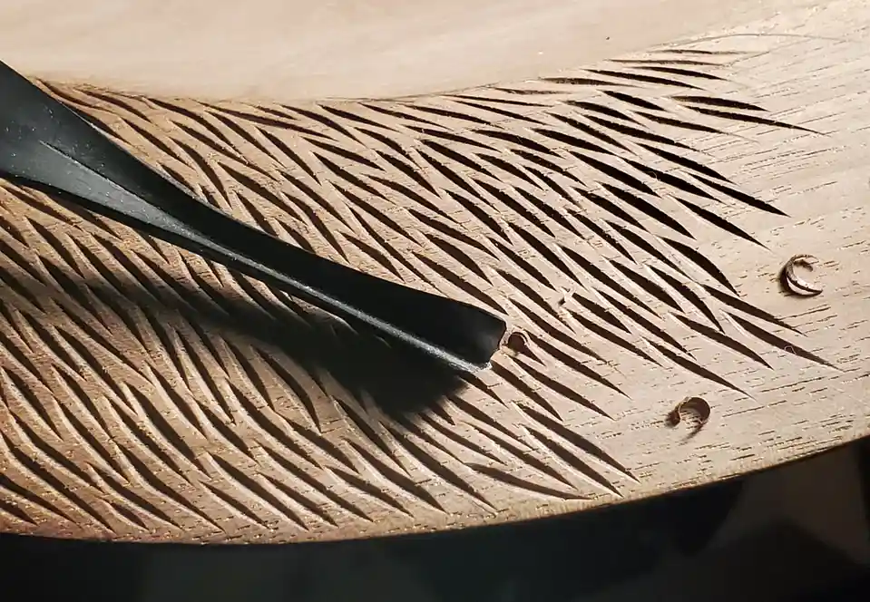 A close-up photo shows a black chisel resting on a wooden surface intricately carved with a feather pattern, featuring fine…