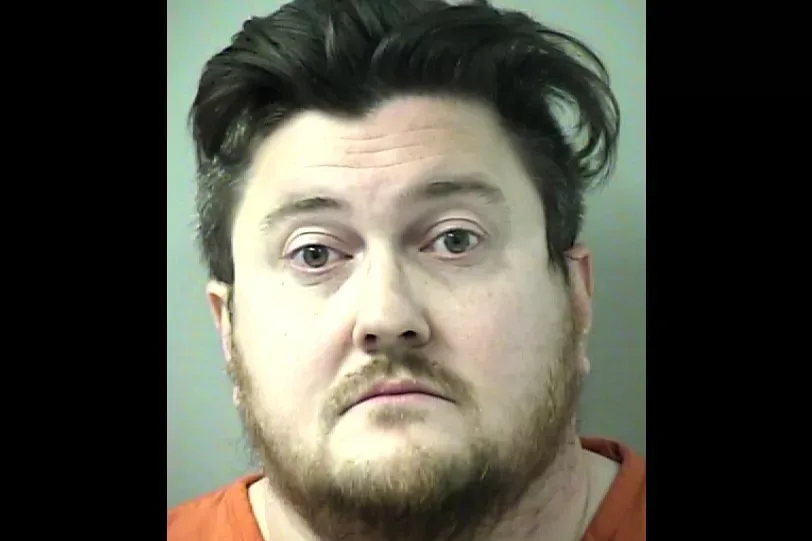 A mugshot of a man with dark hair and a beard wearing an orange shirt, looking directly at the camera with a neutral…