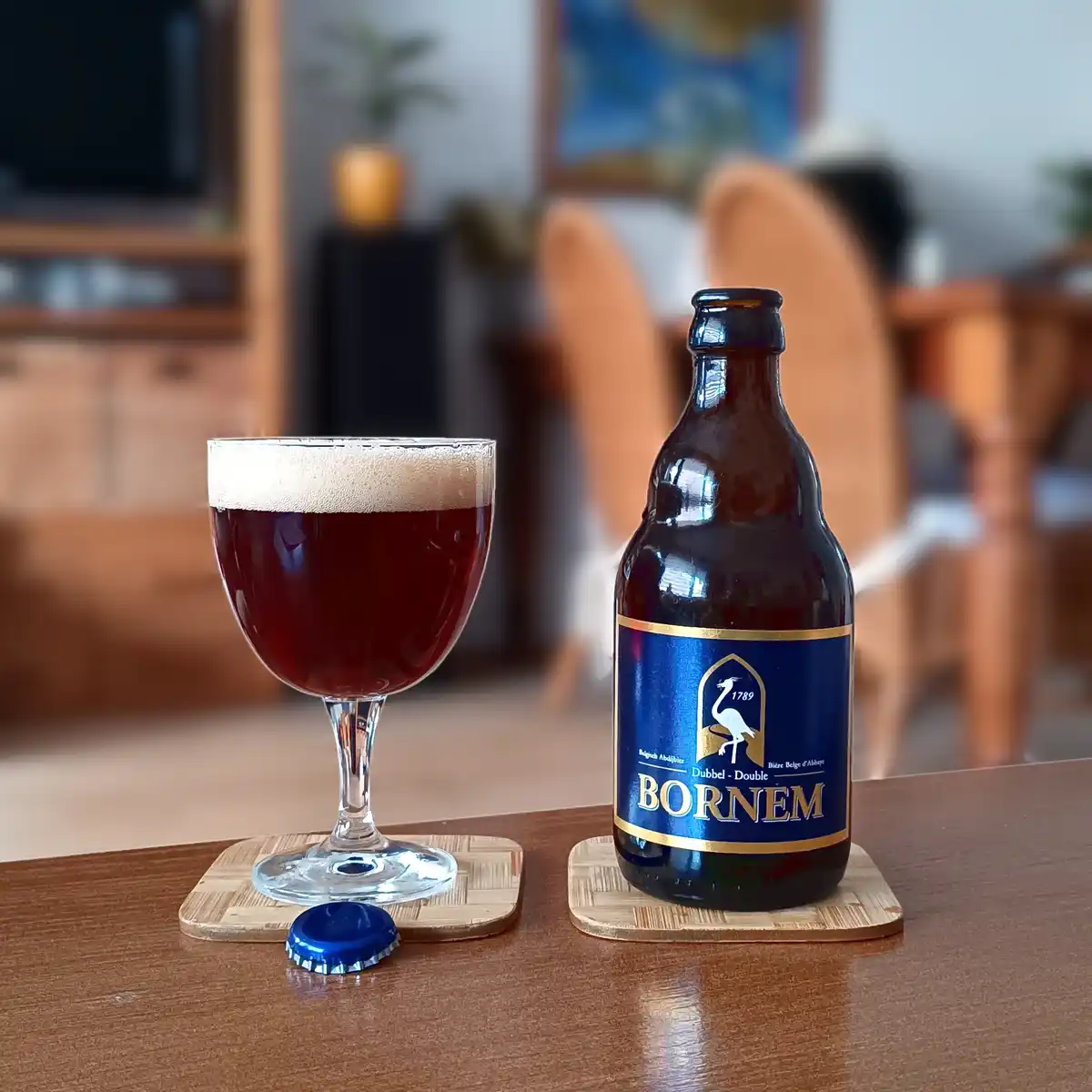 A dark amber beer bottle labeled 'BORNEM' with a blue cap sits next to a filled beer glass on wooden coasters, with a blue…