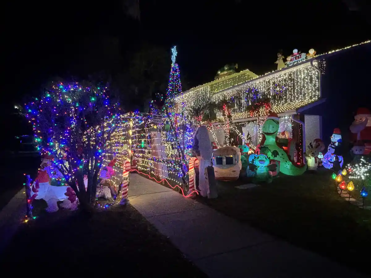 A house at night is extravagantly decorated with Christmas lights, inflatable characters including a large green dinosaur…