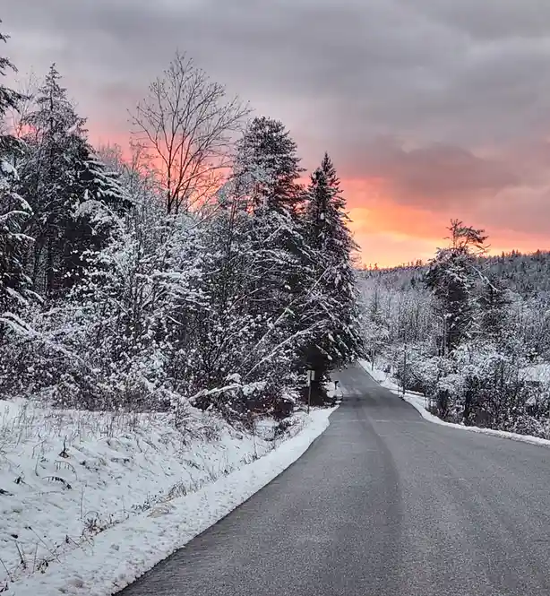 A snowy road winds through a forest at sunset, with trees heavily laden with snow and a dramatic orange-pink sky. Overlaid…