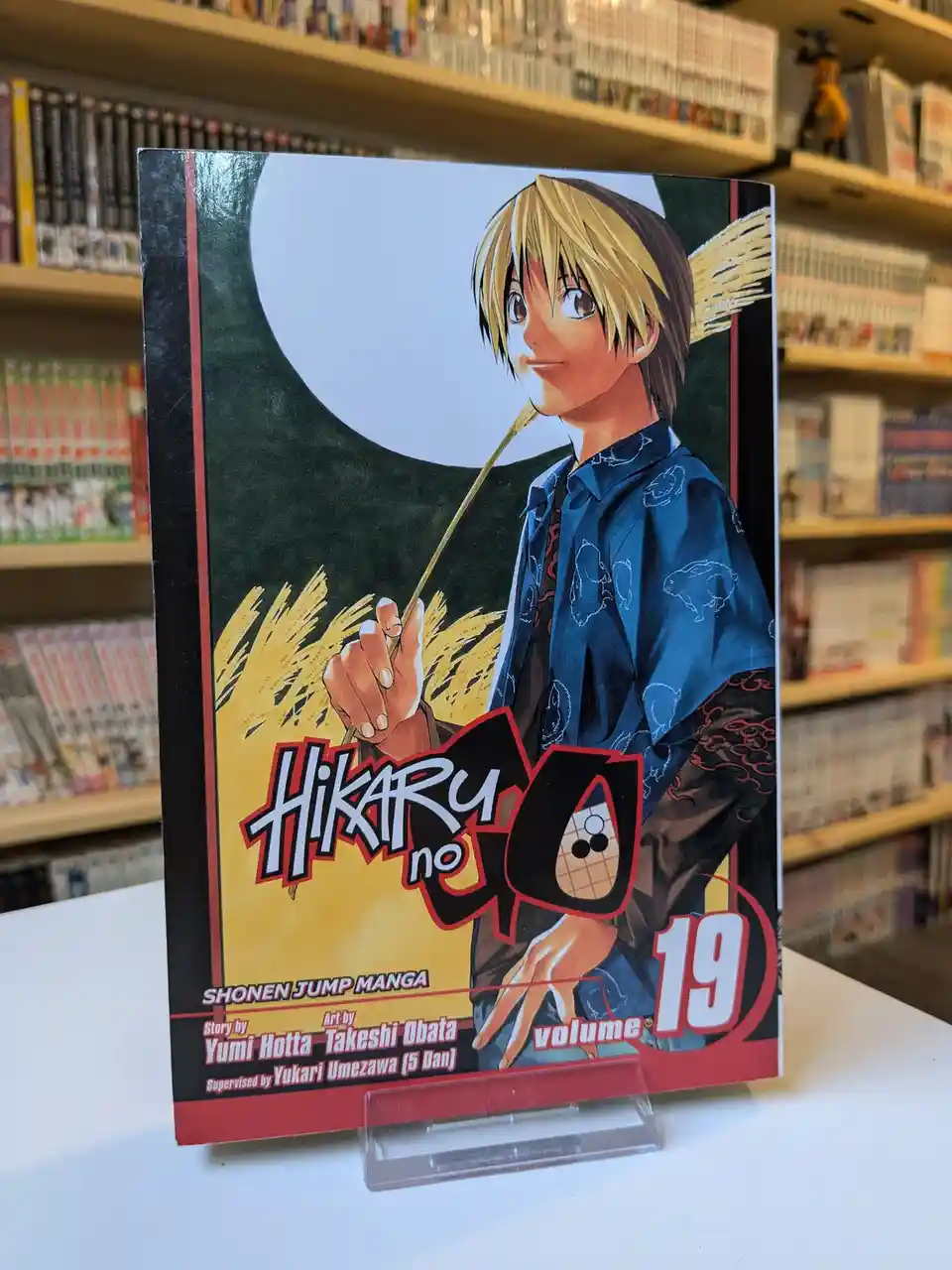 A physical copy of the manga 'Hikaru no Go', volume 19, is displayed upright on a clear stand in a bookstore, with shelves…