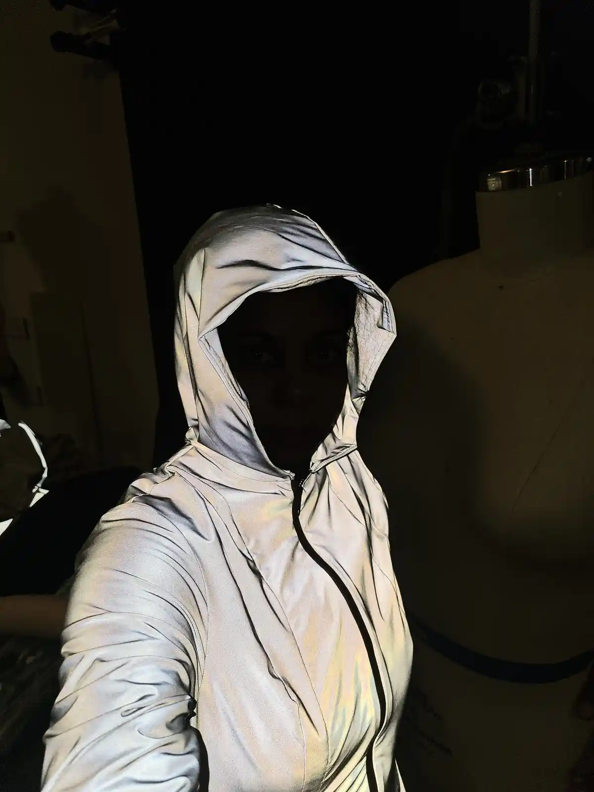 A person wearing a shiny silver reflective hoodie takes a selfie in a dimly lit room, with a mannequin visible in the…