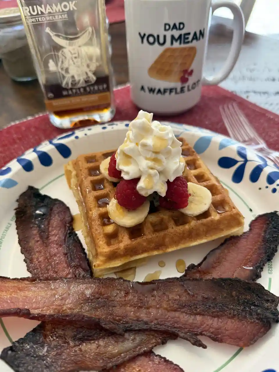 A close-up photo of a breakfast plate with a waffle topped with whipped cream, raspberries, and banana slices, surrounded by…