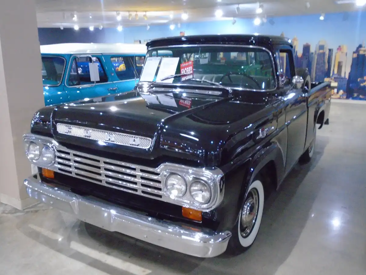 A glossy black vintage Ford pickup truck is displayed indoors, likely in a museum or showroom, with a cityscape mural in the…