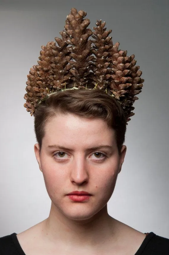 A person with short brown hair wears a crown made of several pinecones arranged on their head, looking directly at the…