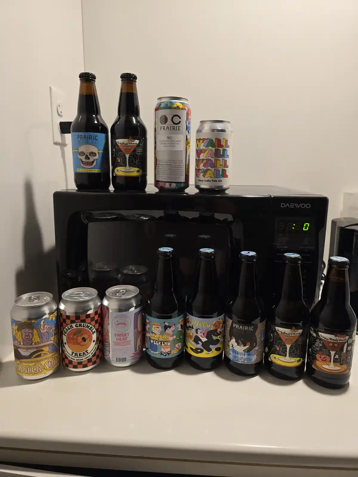 A collection of Prairie Artisan Ales beer bottles and cans are arranged on a white countertop, with several sitting atop a…