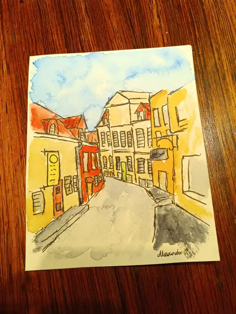 A watercolor painting on paper depicts a European-style street scene with colorful buildings, red roofs, and a gray road…