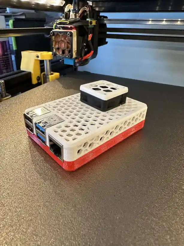 A 3D-printed Raspberry Pi case, white with a red base, sits on a printer bed with a 3D printer's extruder visible above. The…