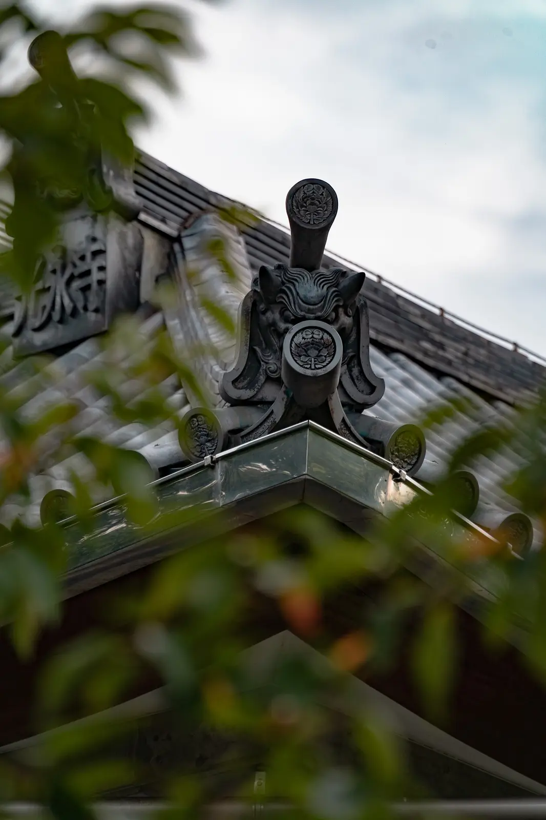A low-angle photograph shows the ornate, dark metal roof ornament of a traditional Japanese temple, featuring a fierce…