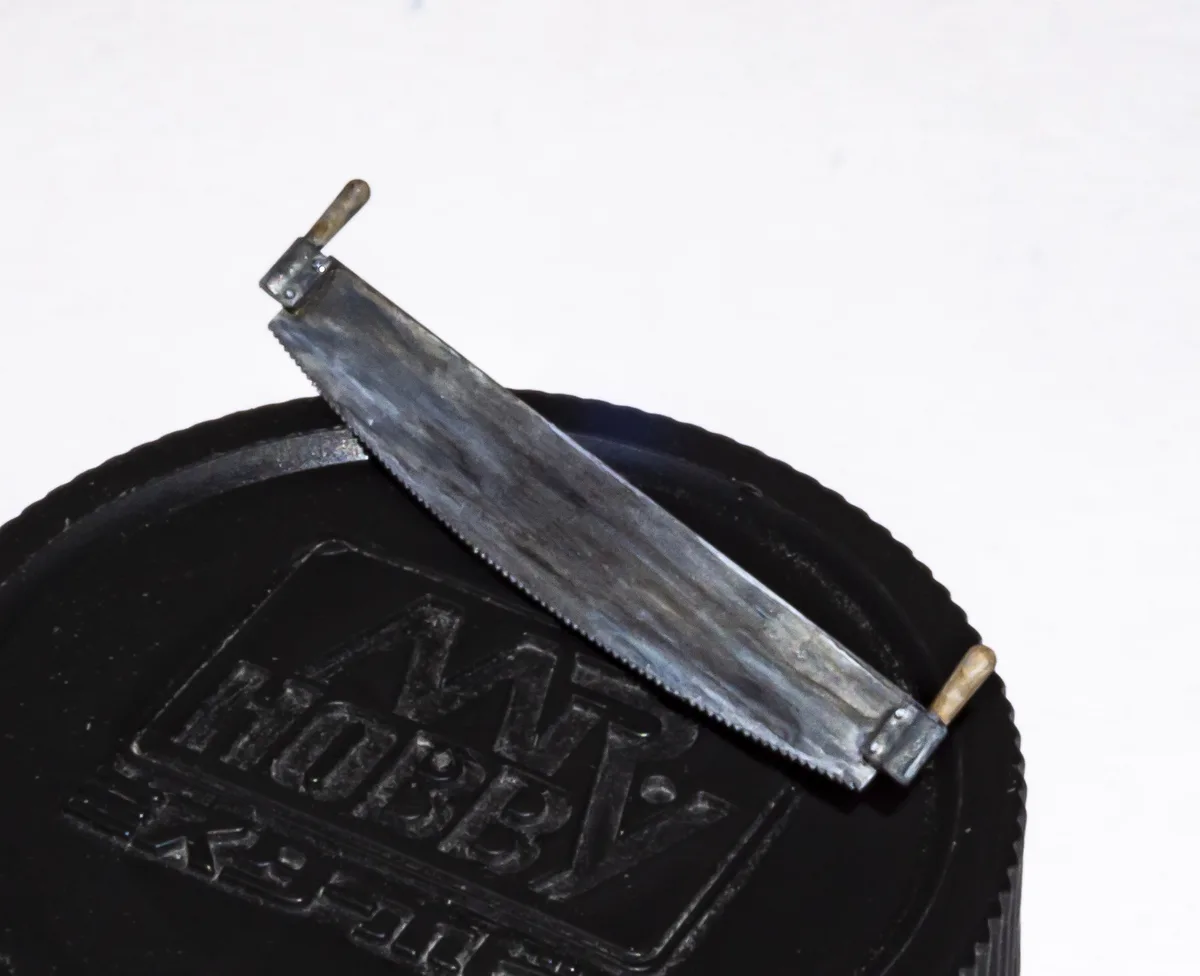 A tiny, handcrafted metal hacksaw rests diagonally across a black circular object embossed with the 'MR HOBBY' logo. The saw…