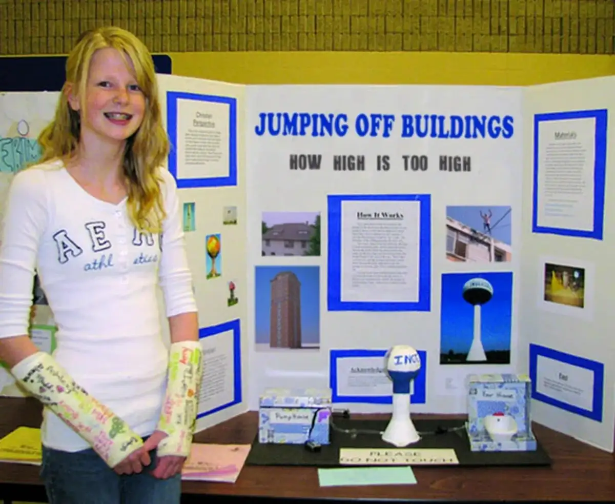 A smiling young girl with both arms in casts stands beside a science fair display board titled 'JUMPING OFF BUILDINGS HOW…