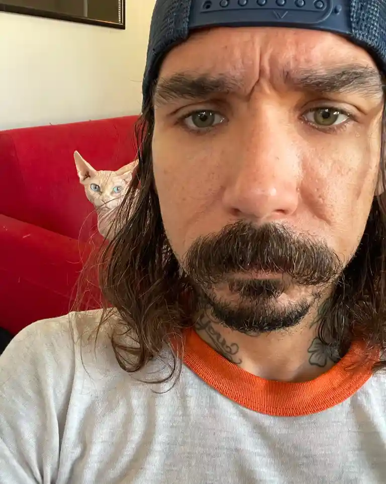 A man with long hair, a mustache, and a backwards cap stares intensely at the camera with a furrowed brow. Behind him, on a…