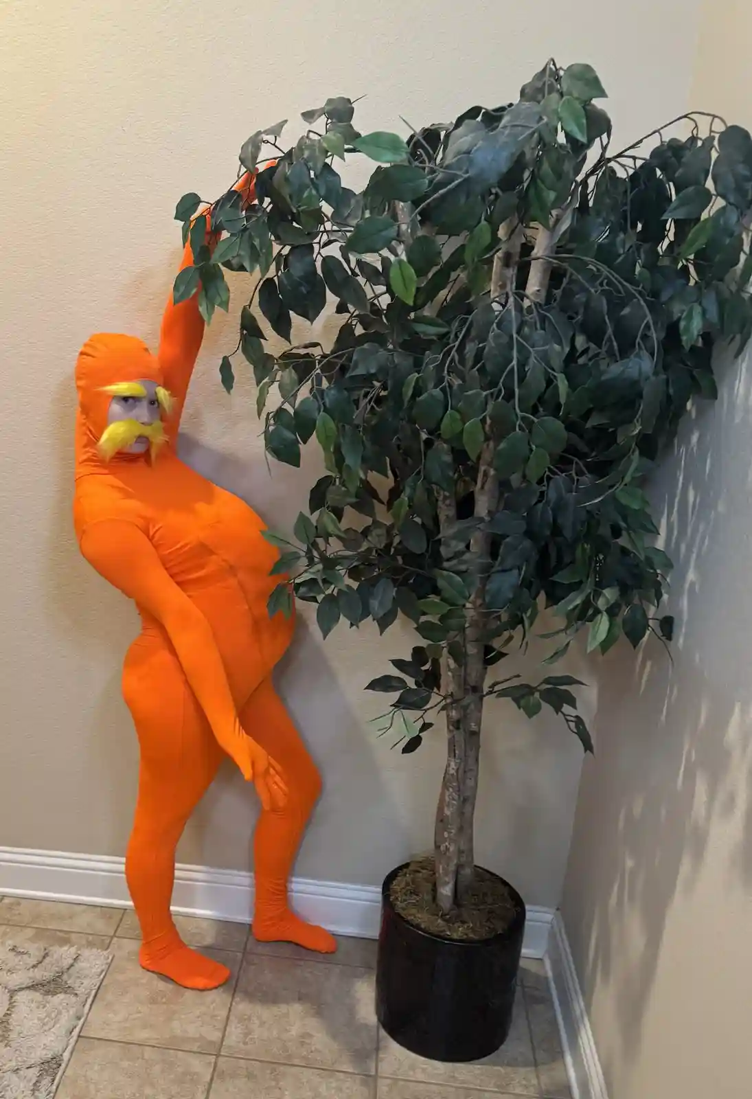 A person in a full-body orange bodysuit with a yellow mustache and eyebrows, mimicking the character Lorax, stands next to a…