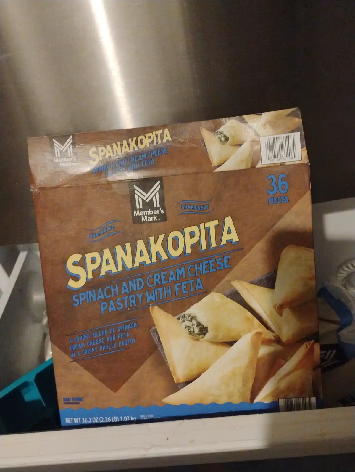 A box of Member's Mark Spanakopita, a spinach and cream cheese pastry with feta, is shown on a shelf. The packaging…