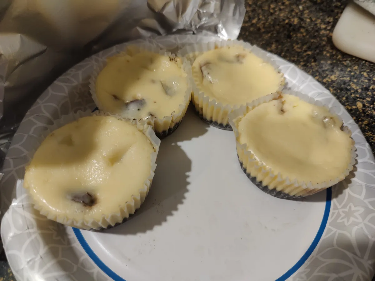 Four unbaked mini cheesecakes in paper liners sit on a patterned paper plate, with a gloved hand visible in the background…