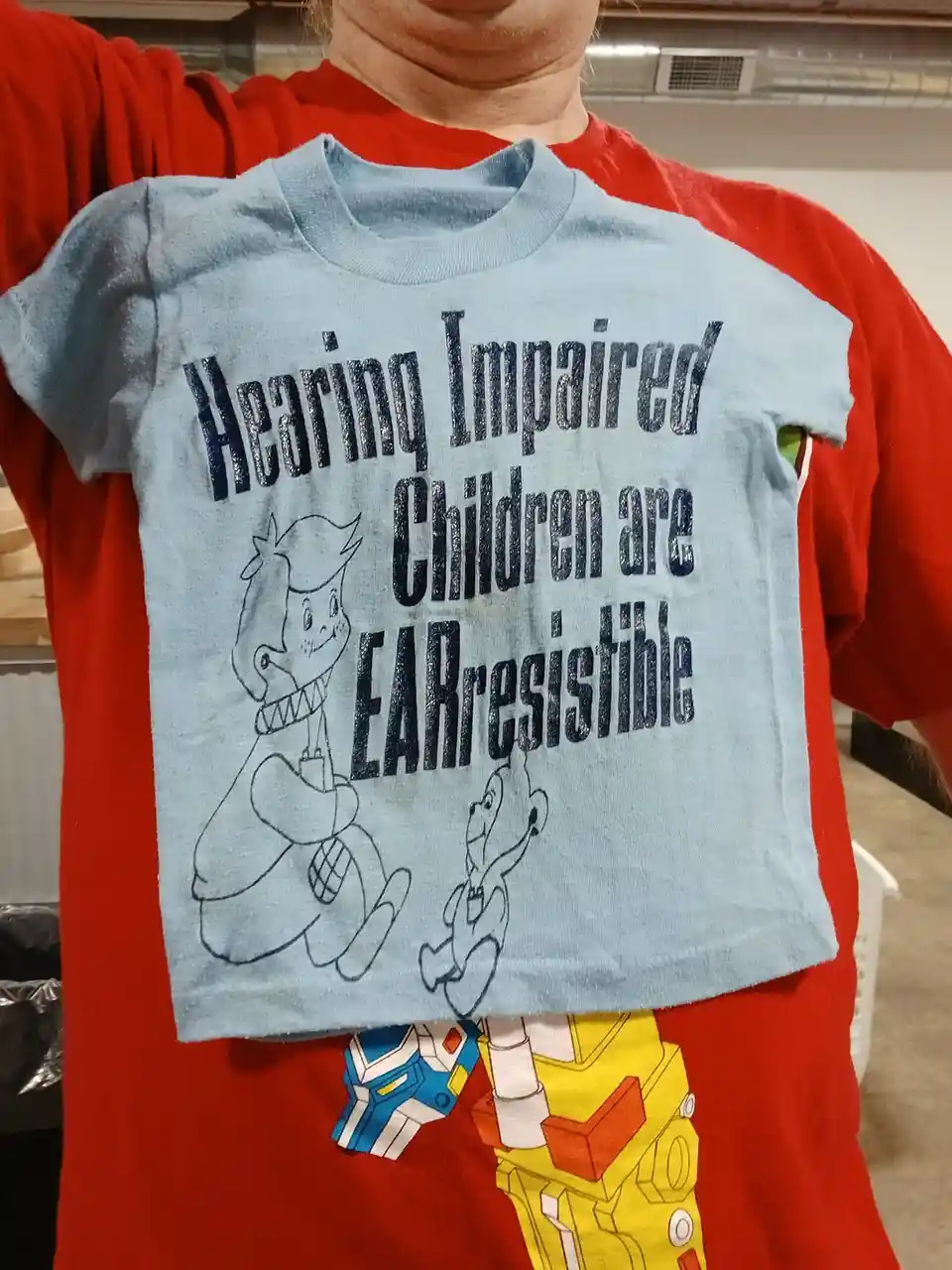 A person holds up a light blue children's t-shirt with black glittery text that reads 'Hearing Impaired Children are…