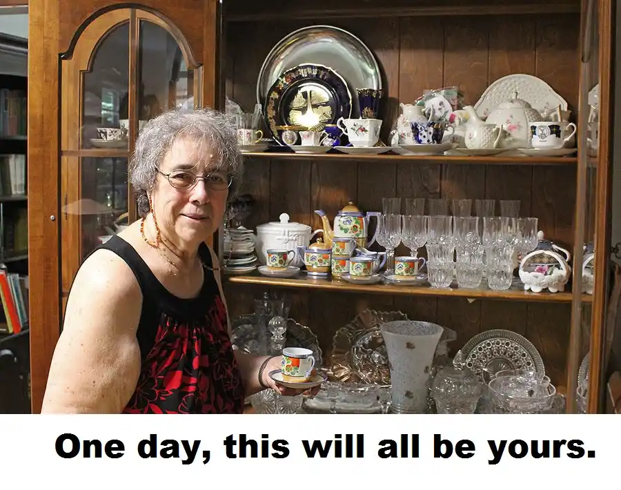 An older woman with glasses and gray hair stands in front of a wooden china cabinet filled with teacups, saucers, glassware…