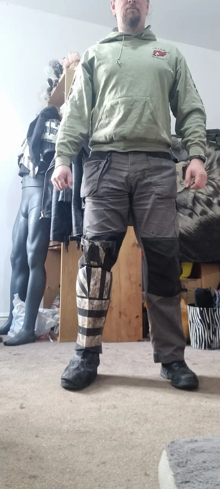 A man stands indoors wearing a green hoodie and cargo pants, with one leg fully covered by a custom-made prosthetic leg…