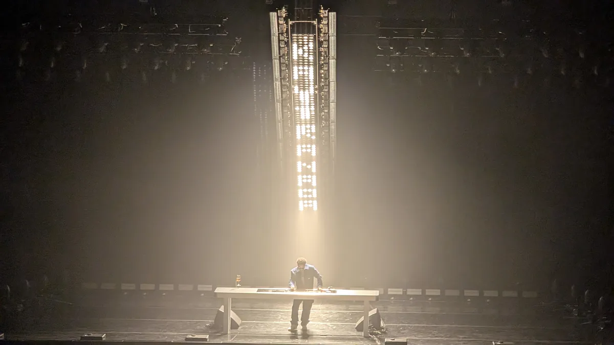A person stands alone on a dimly lit stage behind a long white table, illuminated by a spotlight. Behind them, a tall…