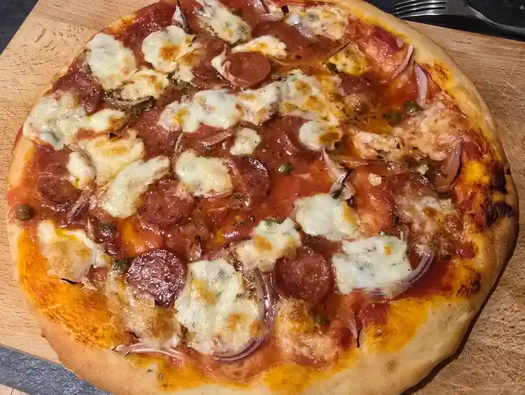 A close-up photo of a homemade pizza with pepperoni, onions, and melted cheese on a wooden cutting board, with a fork…