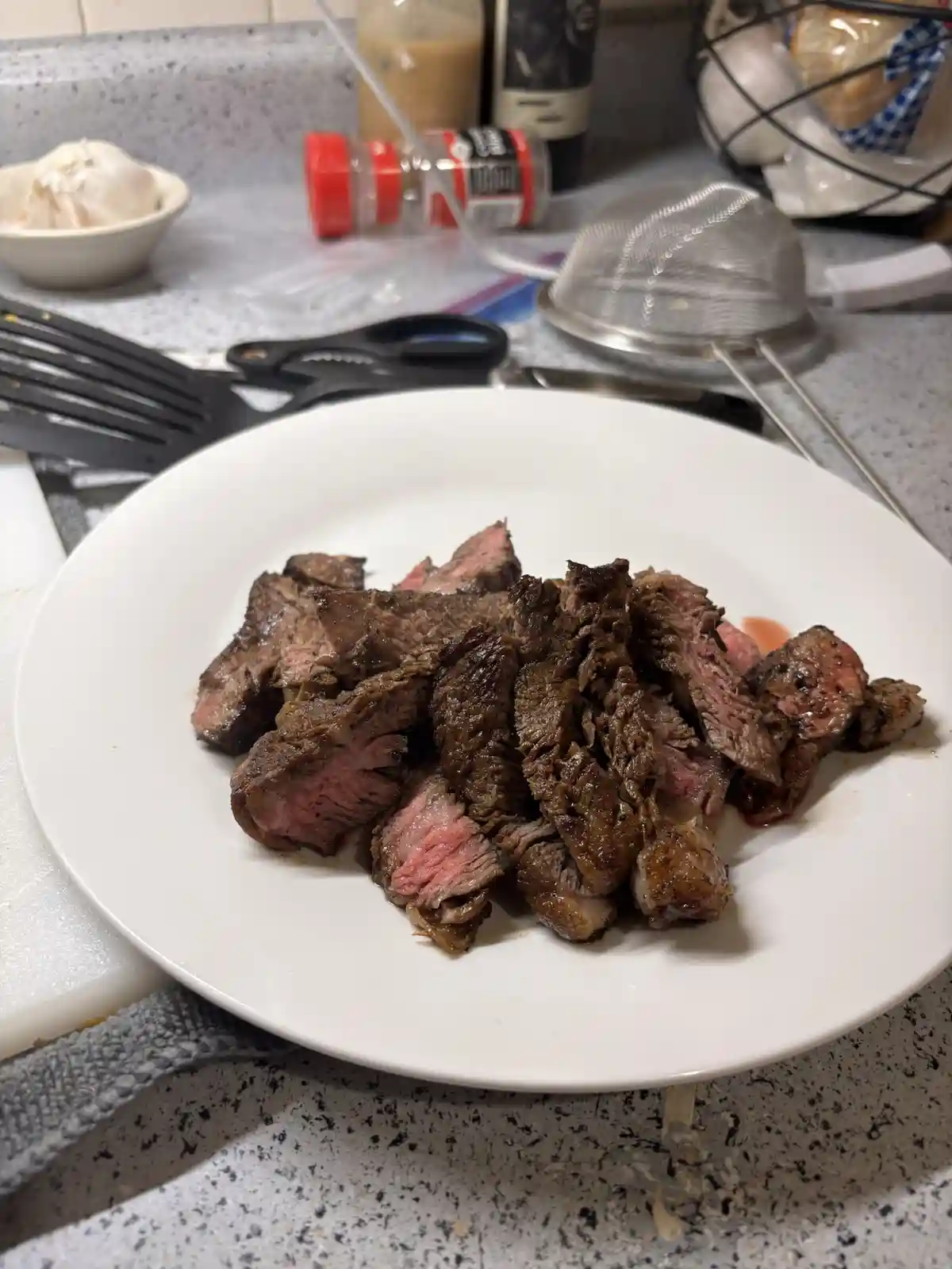 A white plate holds sliced medium-rare steak with pink centers and seared edges, resting on a speckled kitchen countertop…