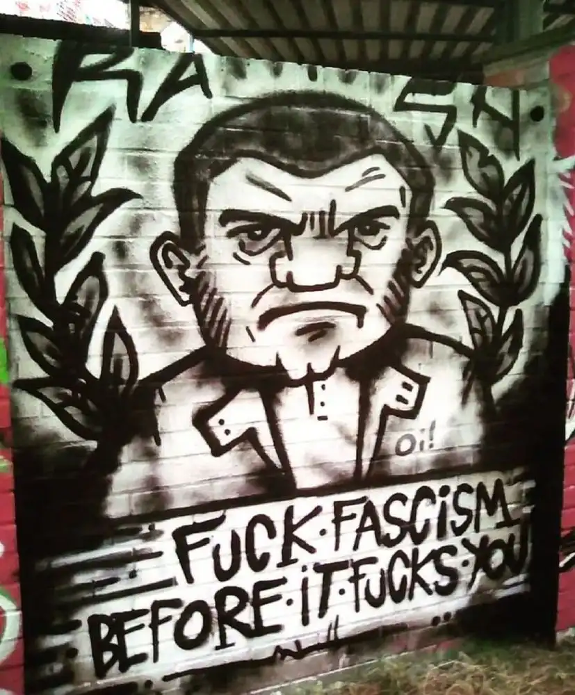 A black-and-white graffiti mural on a brick wall depicts a cartoonish, angry man with a stern expression, framed by laurel…