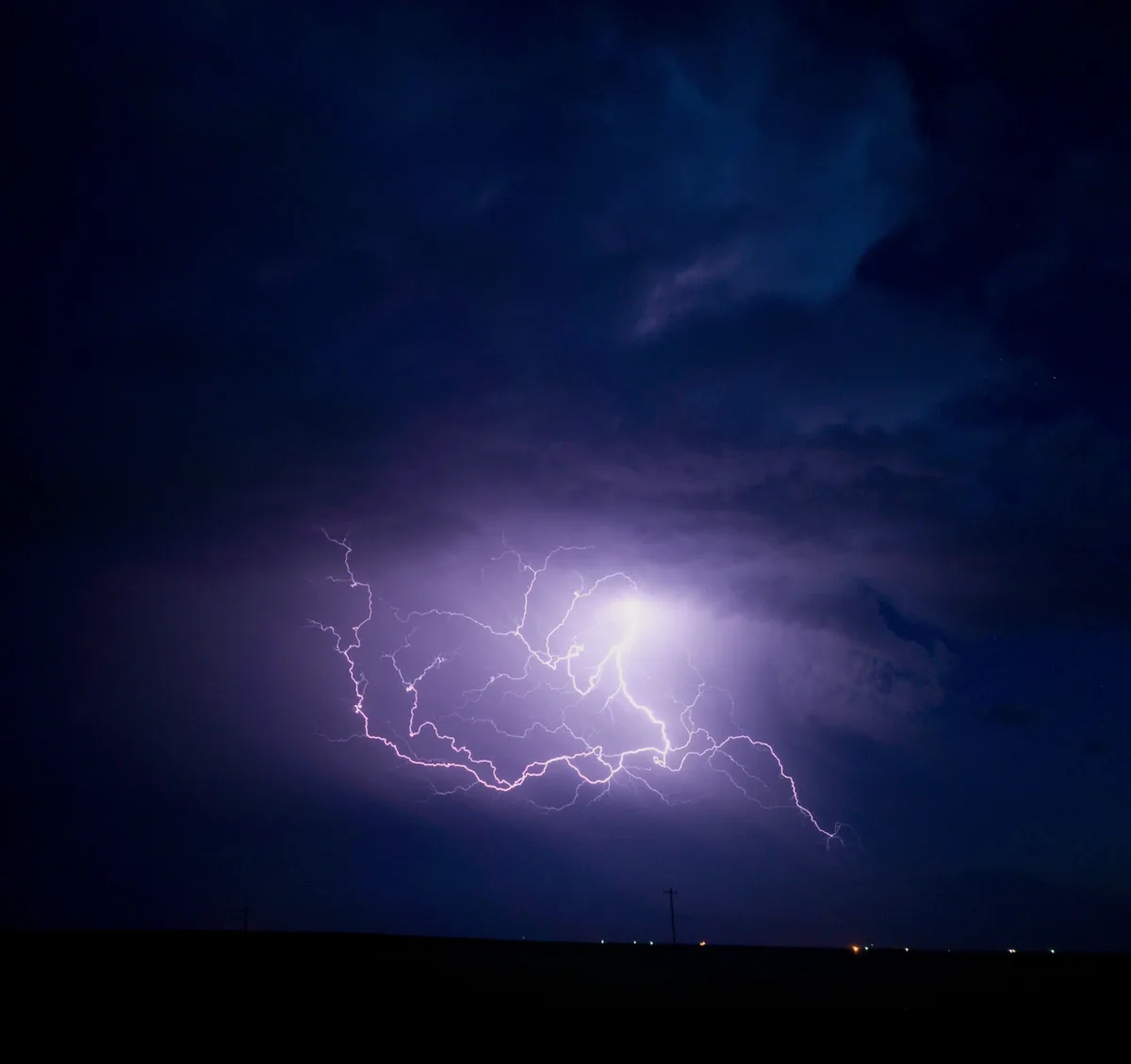 A dramatic nighttime photograph shows a massive, branching lightning bolt illuminating dark storm clouds. The ground below…