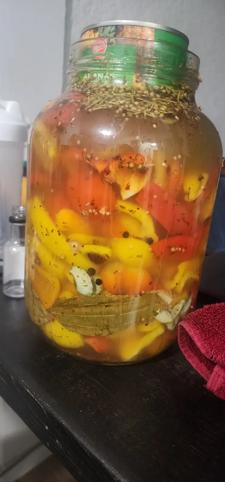 A large glass jar filled with colorful pickled peppers—red, yellow, and orange—soaked in brine with visible spices, bay…