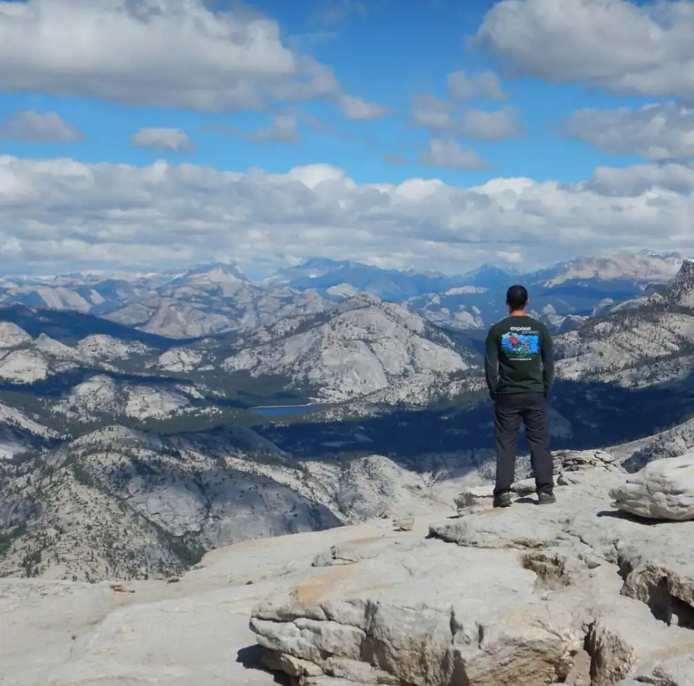 A person stands on a rocky mountain overlook facing away from the camera, gazing at a vast, scenic mountain range under a…