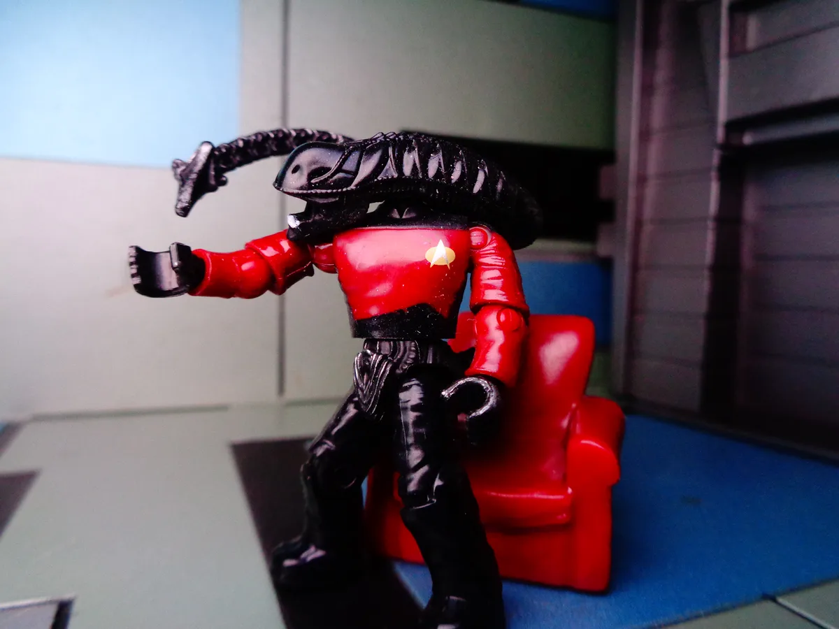 A toy figure with a Starfleet red uniform and gold insignia has an alien creature's head and tail fused to its back…