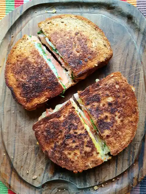Two grilled sandwiches, cut diagonally into four triangular halves, sit on a well-used wooden cutting board. The top…