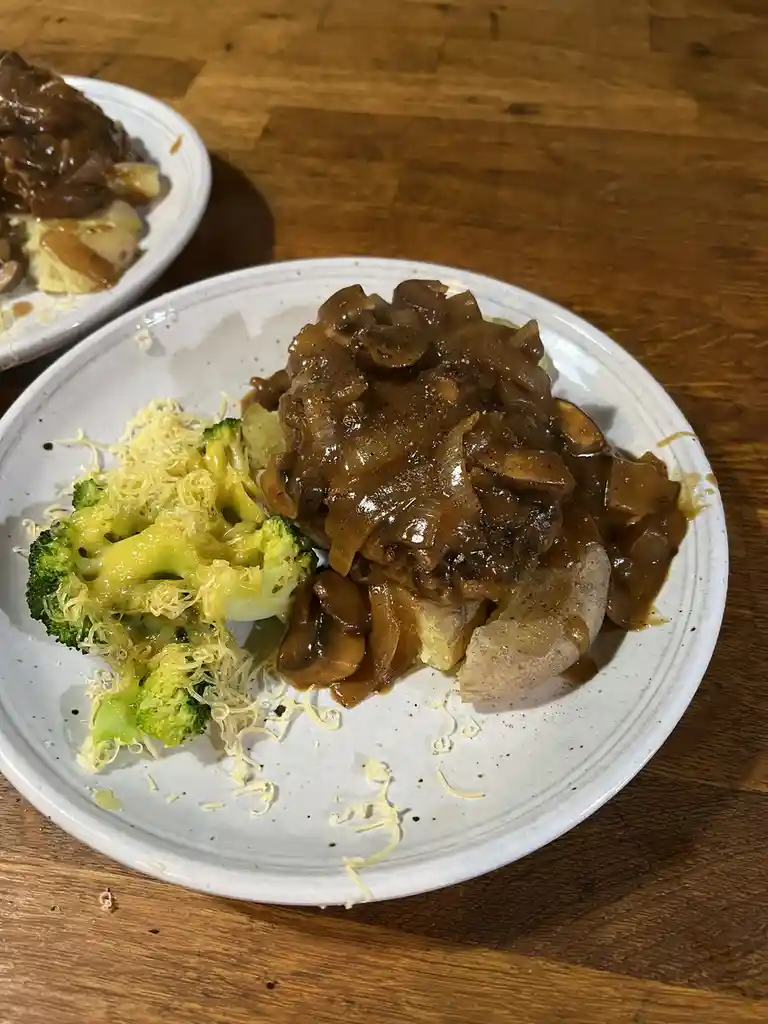 A close-up photo shows a white plate on a wooden table with a steak covered in mushroom gravy, served with a side of…