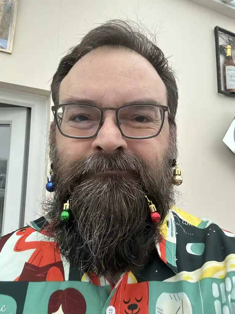 A man with glasses and a full beard is wearing colorful Christmas ornament earrings, standing in front of a wall with a…