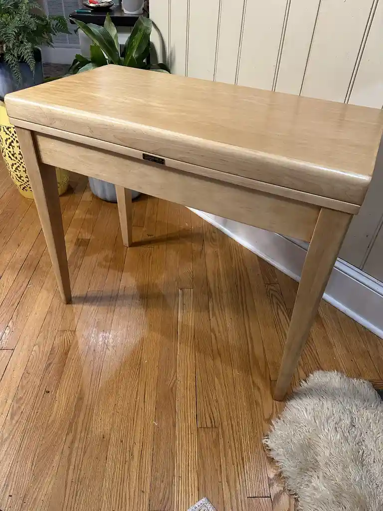 A light-colored wooden piano-style bench with a small plaque reading 'GALLARD BROS INC.' sits on a hardwood floor in a room…