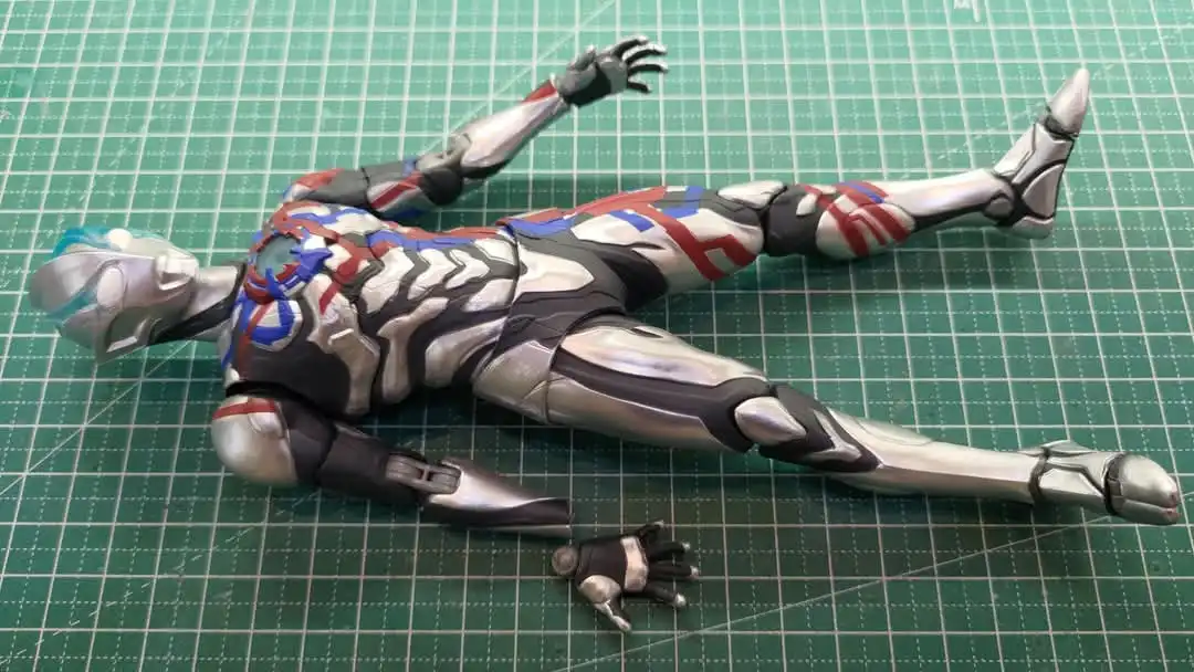 A detailed, silver, red, and blue armored action figure lies on its back on a green cutting mat, one arm raised and one hand…