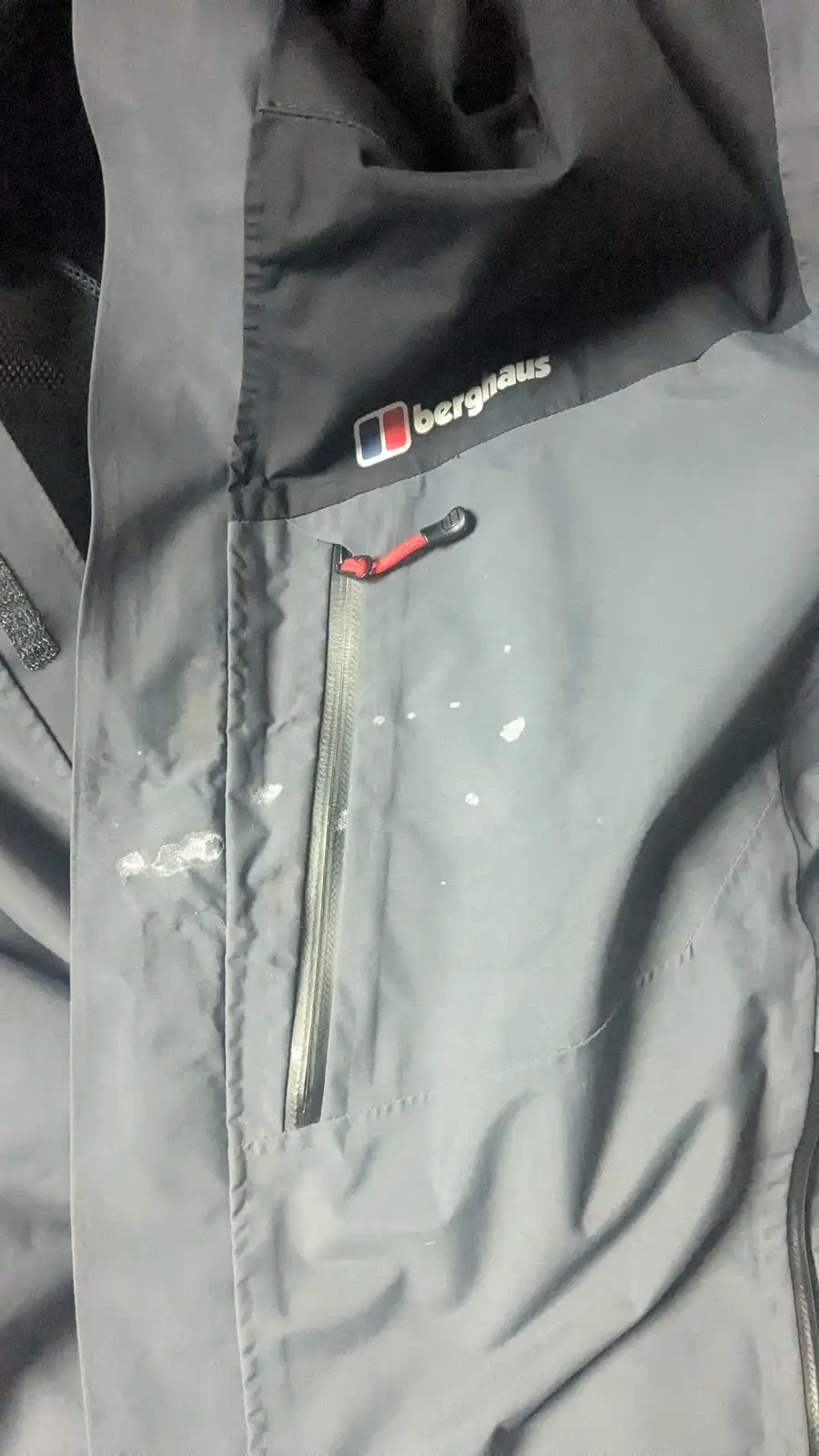 A close-up of a grey Berghaus outdoor jacket with a visible logo and zipper, covered in white splatters resembling paint or…