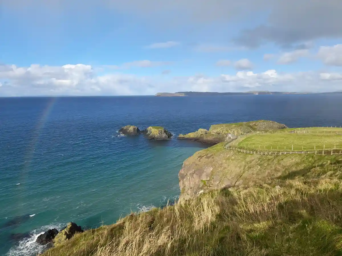 A scenic coastal view shows a faint rainbow arcing over the ocean, with grassy cliffs in the foreground and distant…