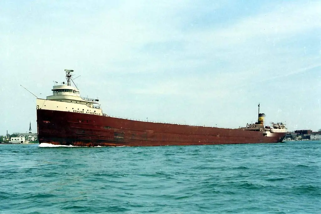 A large, red-hulled freighter named 'EDMUND FITZGERALD' sails on calm blue water under a pale sky, with a distant shoreline…