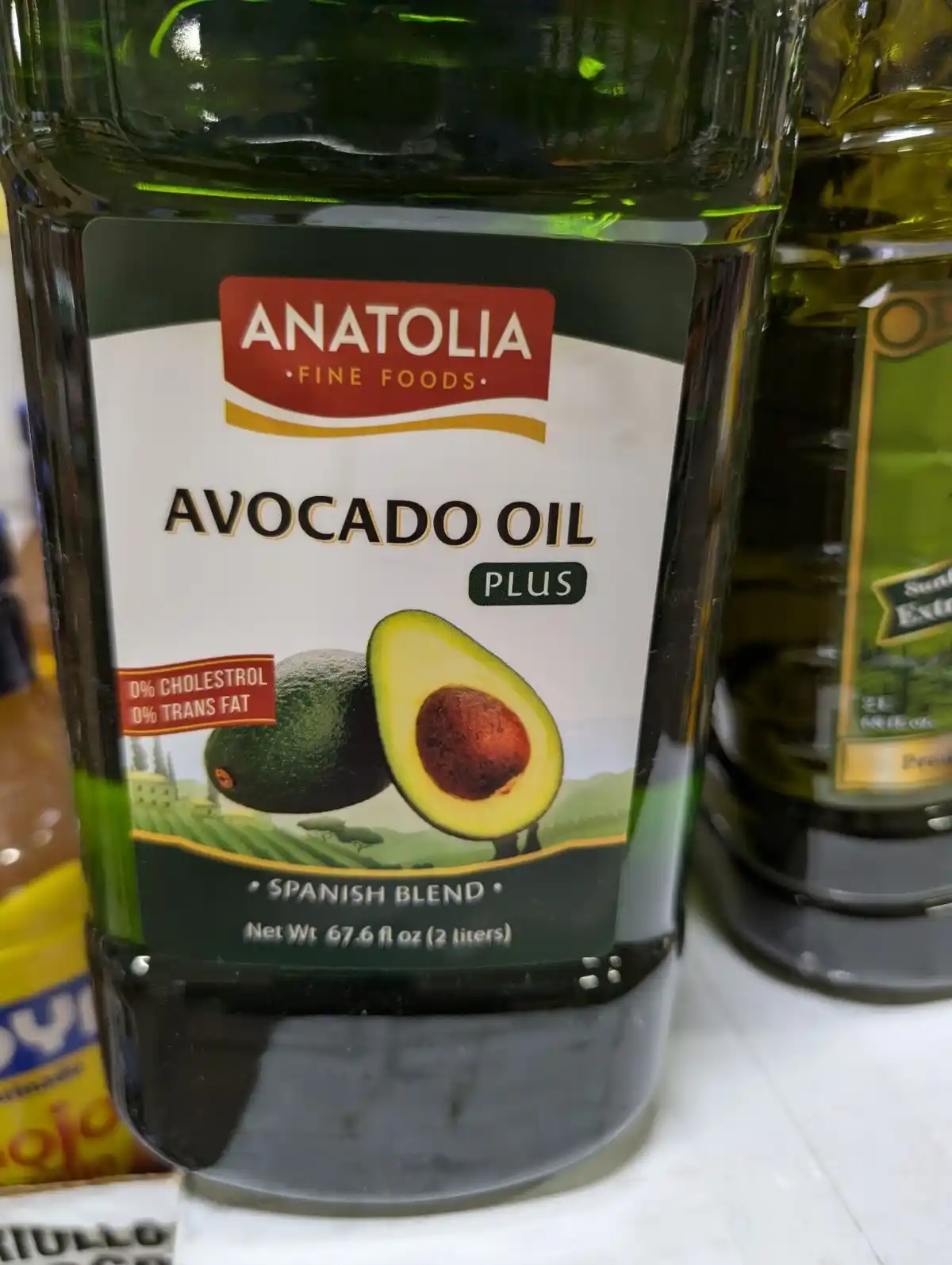 A close-up photo of a large bottle of Anatolia Fine Foods Avocado Oil Plus, featuring an image of whole and halved avocados…