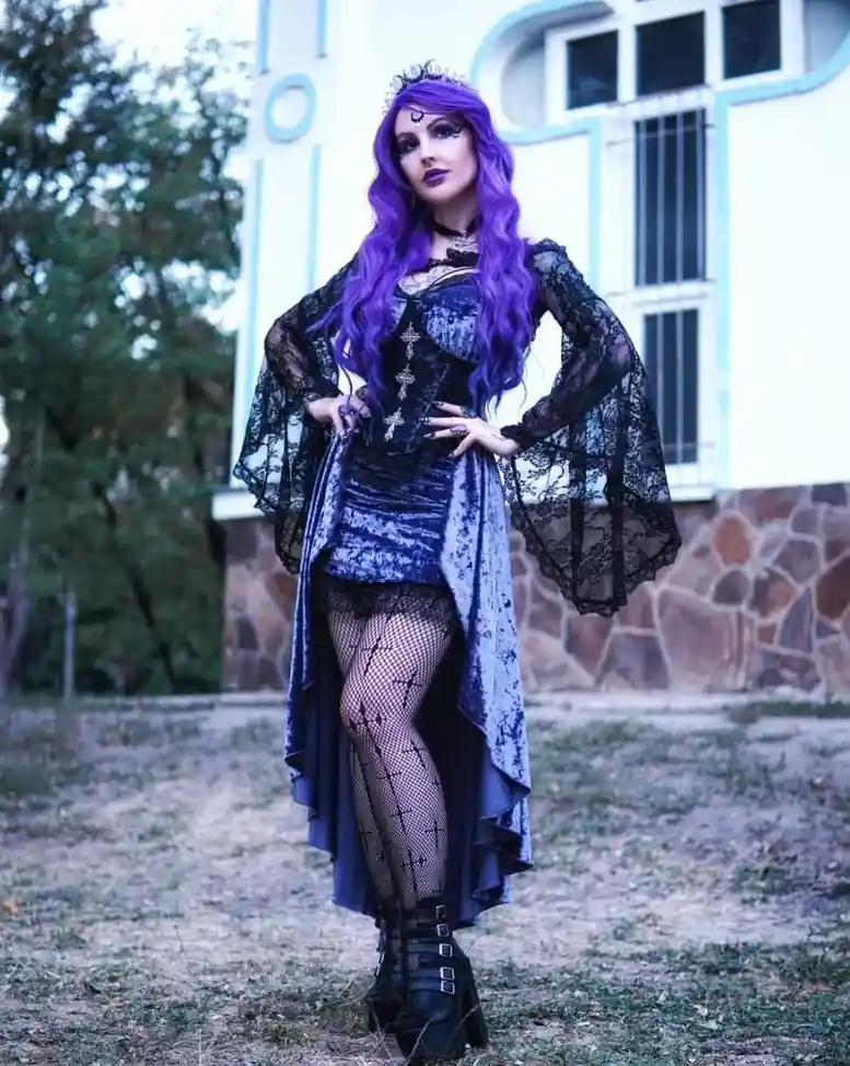 A woman with long purple hair and dark makeup poses confidently outdoors in front of a building. She wears a gothic outfit…