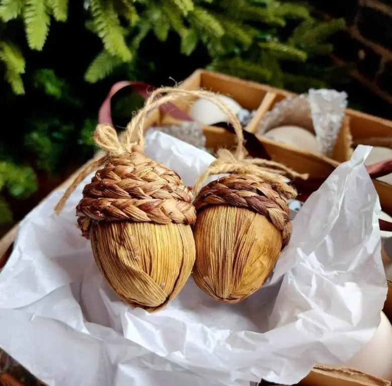 Two handcrafted, woven acorn-shaped ornaments with braided caps and twine loops sit nestled in white tissue paper inside a…