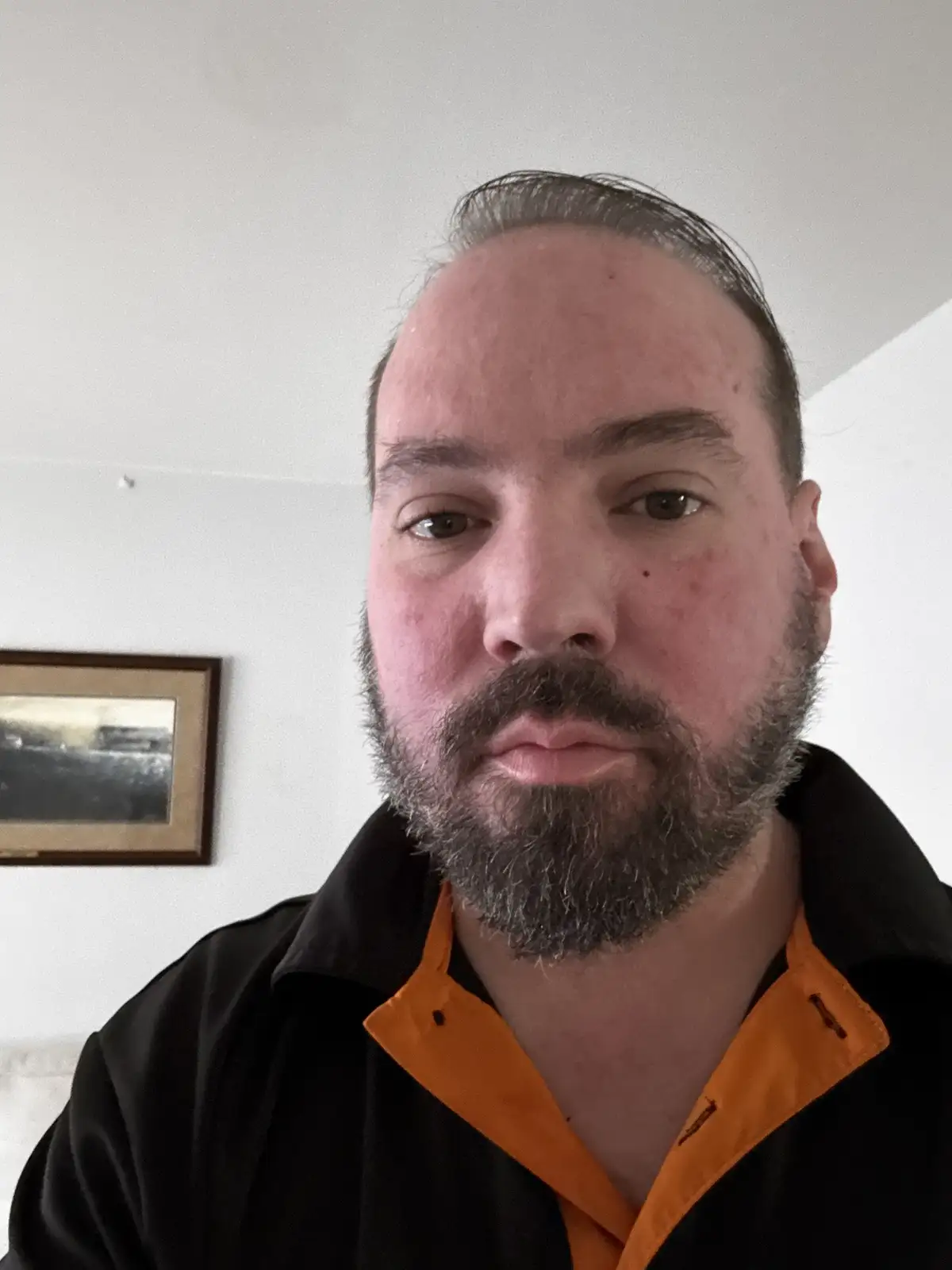 A close-up selfie of a man with a receding hairline and a graying beard, wearing a black shirt with an orange collar…