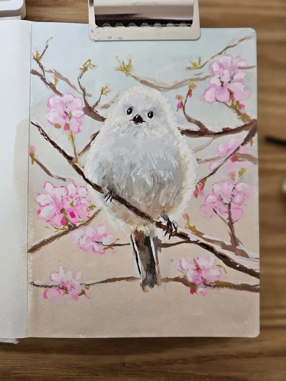 A hand-painted illustration on a sketchbook page shows a fluffy white bird perched on a branch with pink cherry blossoms…