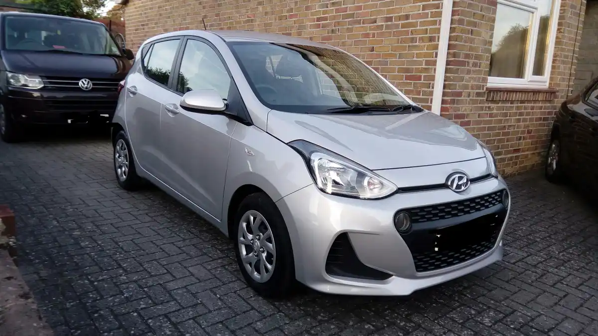 A silver Hyundai i10 is parked on a brick driveway in front of a brick house, with a dark Volkswagen van visible behind it…