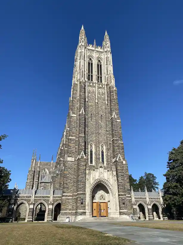 A low-angle photo shows a tall, ornate Gothic-style stone bell tower against a clear blue sky, with trees and a paved…