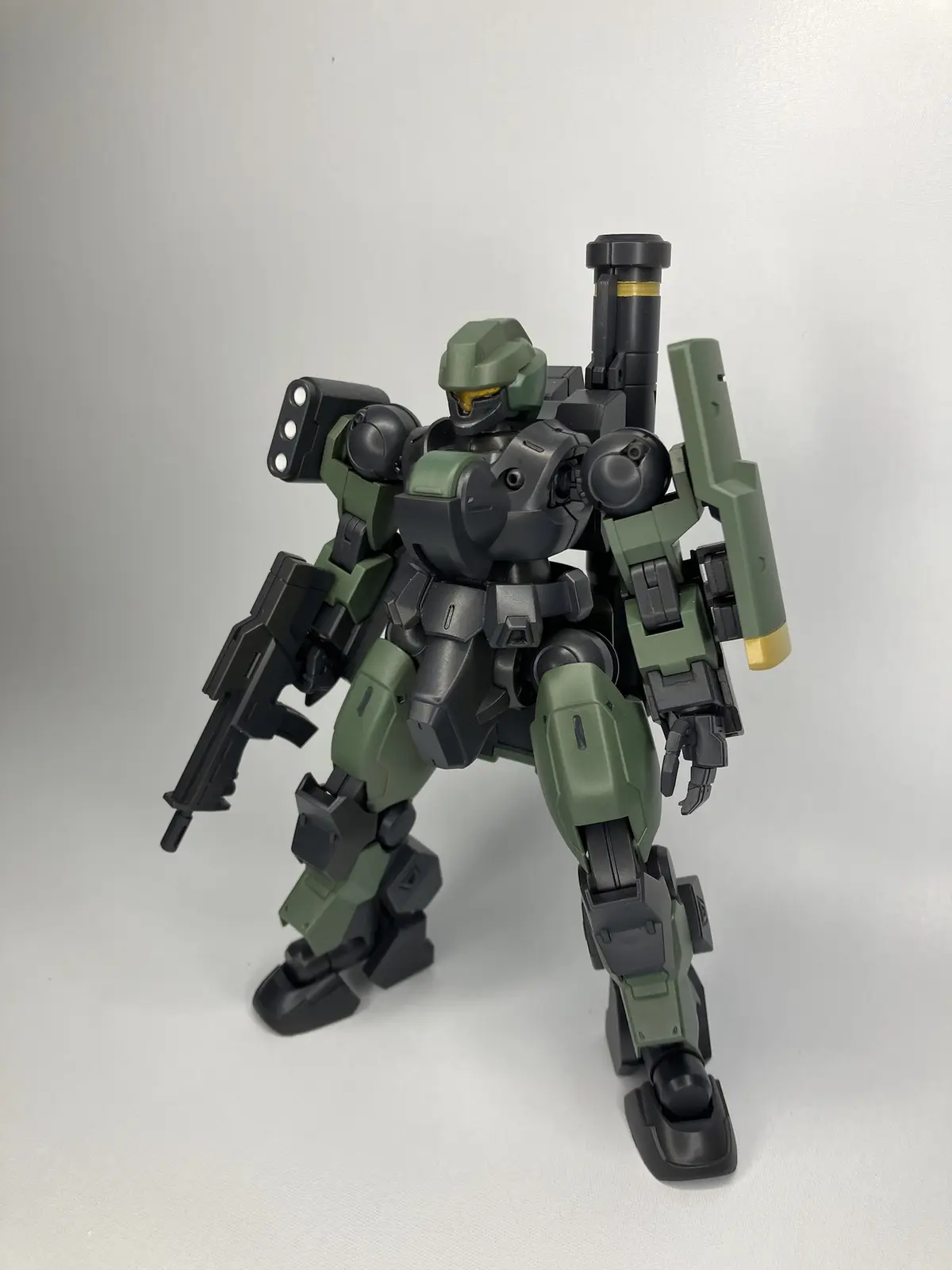 A green and black robot model stands in a combat-ready pose against a plain white background, holding a rifle in one hand…