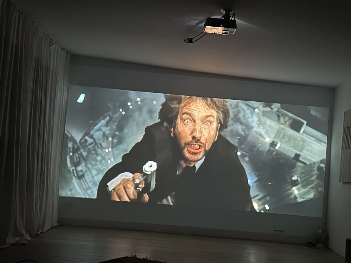 A large projection screen in a room displays a close-up of a man with wild hair and an intense, screaming expression…